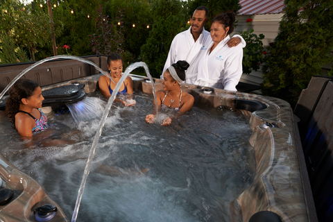 A family in a Premium Collection spa at dusk — children in the water, parents in robes alongside, fountain jets running